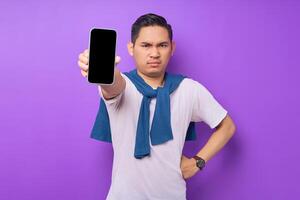 Angry young Asian man holding smartphone with blank screen isolated on purple background. Advertising new mobile app, mockup concept photo