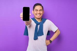 Smiling young Asian man holding smartphone with blank screen isolated on purple background. Advertising new mobile app, mockup concept photo