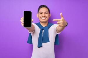Cheerful young Asian man pointing index finger at empty smartphone screen isolated on purple background. Advertising new mobile app, mockup concept photo