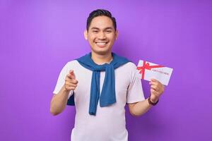 Smiling young Asian man in casual clothes holding gift certificate and pointing finger the camera isolated on purple studio background photo