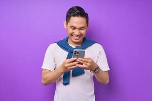 Cheerful young Asian man using smartphone for messaging with friends isolated over purple background photo