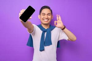 Cheerful young Asian man holding smartphone with blank screen and gesturing okay sign isolated on purple background. Advertising new mobile app, mockup concept photo