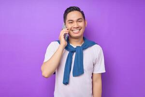 Cheerful young Asian man in white t-shirt talking on mobile phone isolated on purple background photo