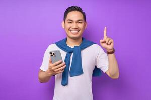Portrait of cheerful handsome young in casual clothes holding smartphone having an idea and pointing upwards isolated on purple background photo