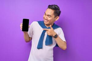 Cheerful young Asian man pointing index finger at empty smartphone screen isolated on purple background. Advertising new mobile app, mockup concept photo