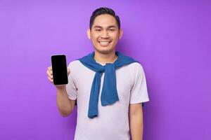 Smiling young Asian man wearing casual clothes showing mobile phone with blank screen isolated on purple background. Advertising new mobile app, mockup concept photo