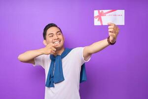 Smiling young Asian man in casual clothes pointing a finger at gift voucher certificate and looking at the camera isolated on purple studio background photo