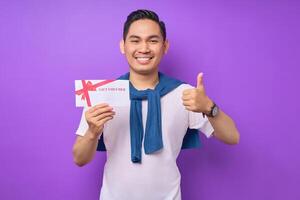 Smiling young Asian man in casual clothes holding gift voucher certificate and showing thumb up isolated on purple studio background photo