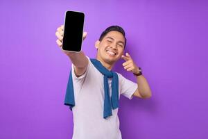 Cheerful young Asian man pointing index finger at empty smartphone screen isolated on purple background. Advertising new mobile app, mockup concept photo