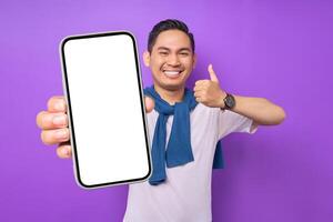 Cheerful young Asian man demonstrating smartphone with empty screen, showing thumb up gesture isolated on purple background. Advertising new mobile app, mockup concept photo