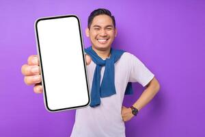 Smiling young Asian man holding smartphone with blank screen isolated on purple background. Advertising new mobile app, mockup concept photo