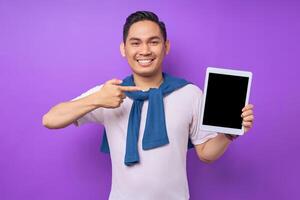 Smiling young Asian man in casual clothes pointing finger at digital tablet with empty screen for mockup isolated over purple background photo
