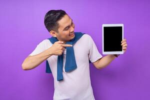 Smiling young Asian man in casual clothes pointing finger at digital tablet with empty screen for mockup isolated over purple background photo