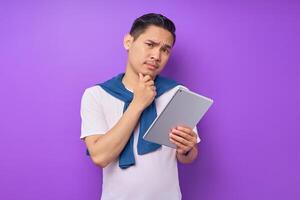 Portrait of pensive young Asian man in white t-shirt touching chin and holding digital tablet isolated on purple background photo