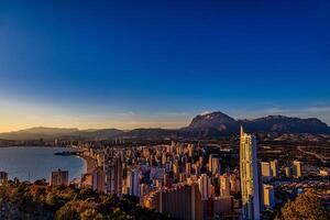 A coastal cityscape Benidorm is bathed in the warm glow of a setting sun, with high-rise buildings clustering along the shoreline photo