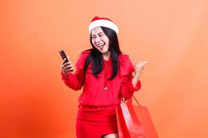 portrait of Asian girl wearing christmas blouse, Santa hat, happy to mobile phone, hand sign success holding device, carrying red bag of christmas gifts, isolated on orange background photo