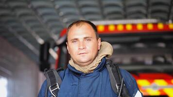 Portrait of tired male firefighter standing near a fire engine at department. Young exhausted fireman in protective uniform looking at camera after fire against the background of big red truck photo