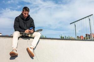 copyspace Relaxed skater texting on smartphone while sitting at half-pipe edge during break photo
