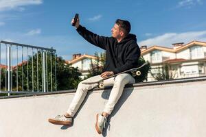 Skater taking a selfie with smartphone while sitting on half-pipe edge in skate park photo