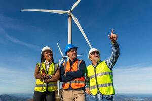 three multiethnic Engineers pointing at wind turbine in wind farm generating green energy photo