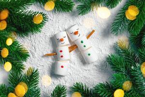 Two adorable marshmallow snowmen are embracing on a snowy background surrounded by christmas tree branches and warm, inviting lights, creating a festive and heartwarming scene photo
