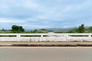 Horizontal view of concrete road across river, side of concrete bridge fence, background is green forest and mountains under sky with river flowing through. At Mae Ngao Mae Hong Son of Thailand. photo