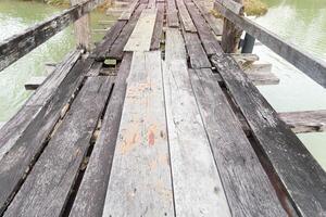 Weathered wooden bridge with some planks missing and others cracked and uneven. Spans a body of water. The water below reflects the cloudy sky and the bridge worn surface. photo