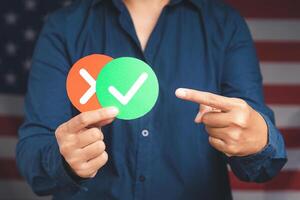 Close-up of hand holding two papers with a checkmark and cross while standing on the American flag background photo