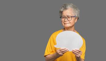 Senior woman holding a blank speech bubble and looks at the camera with a smile while standing on a gray background photo