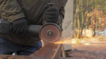 A worker wearing gloves operates a power grinder on a metal beam, creating sparks in a tranquil wooded environment during the day, showcasing craftsmanship and focus. video