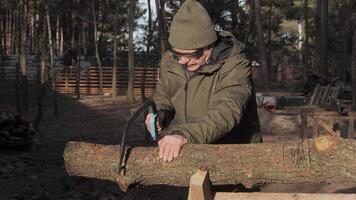 A man wearing a green jacket and beanie is sawing a log in a wooded area. Sunlight filters through the trees, creating a peaceful and focused atmosphere for this task. video