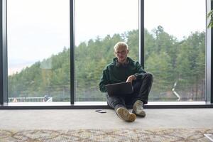 A blonde man sitting on the floor of a modern office, leaning against the window with a laptop in his lap, fully engaged in his work while maintaining a relaxed and focused demeanor. photo