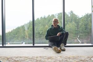 A blonde man sitting on the floor of a modern office, leaning against the window with a laptop in his lap, fully engaged in his work while maintaining a relaxed and focused demeanor. photo