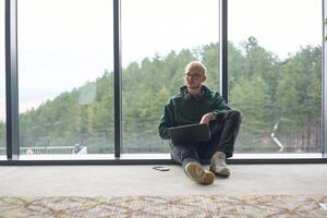 A blonde man sitting on the floor of a modern office, leaning against the window with a laptop in his lap, fully engaged in his work while maintaining a relaxed and focused demeanor. photo