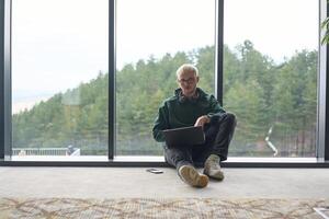 A blonde man sitting on the floor of a modern office, leaning against the window with a laptop in his lap, fully engaged in his work while maintaining a relaxed and focused demeanor. photo