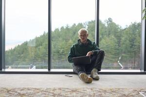 A blonde man sitting on the floor of a modern office, leaning against the window with a laptop in his lap, fully engaged in his work while maintaining a relaxed and focused demeanor. photo