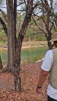 4k footage of an angler walking among trees with fallen leaves around a river flowing. An adult Asian man is walking in slow motion along the river looking for a fishing spot on the river bank. video