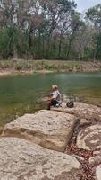 The activity of fishing alone on the edge of a calm and quiet river. 4k footage of an angler fishing waiting for his bait to be eaten by fish on the edge of a clear river full of rocks. video