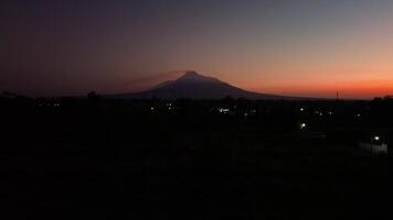 4k aerial footage of Mount Merapi from a distance when it was still dark before sunrise. The panorama towards the morning of the active volcano from a distance is still a silhouette with a thin light video