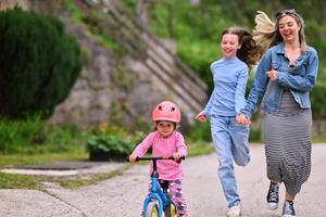 A cheerful little girl riding a balance bike with a helmet while two women joyfully run beside her on a paved path in a lush outdoor setting. photo