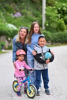 A happy group of people, including two women, a boy holding a ball, and a little girl on a balance bike with a helmet, enjoying outdoor activities on a paved path surrounded by greenery. photo