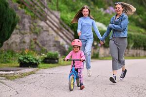 A cheerful little girl riding a balance bike with a helmet while two women joyfully run beside her on a paved path in a lush outdoor setting. photo