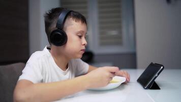 Cute boy looking on a tablet while eating. Side view of a little boy in headphones watching on a wireless gadget and eats slowly. video