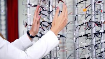 Young female choosing eyeglasses in the store. Hands of a woman customer spinning the stand with fashionable spectacles at optics. video