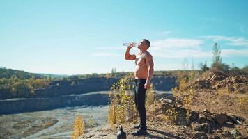 Side view of a sportsman drinks water after workout. Young athlete standing on the rocky hill and drinking water from a bottle in summer. video