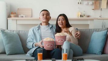 A young couple sits on a cozy sofa, sharing a fun movie night. They hold large popcorn buckets and have refreshing drinks beside them while watching television together. video