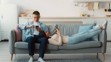 A couple is relaxing together on a sofa in a bright living room. The man sits upright, focused on his phone, while the woman lies back, also engaged with her device. video