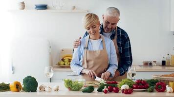 A couple enjoys cooking together in a warm kitchen filled with fresh vegetables and herbs. The woman chops ingredients while the man stands behind her, highlighting their teamwork and affection. video
