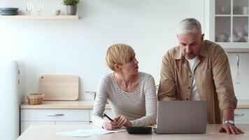 Two adults focus on a laptop in a well-lit kitchen, collaborating on a task while reviewing documents. The setting is organized with minimalistic decor and a cozy atmosphere. video