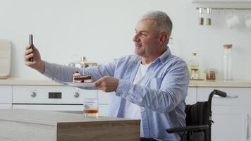 A middle aged Caucasian man uses his smartphone to take a selfie while holding a cake with a lit candle. He is seated in a wheelchair at a kitchen table in his home. video
